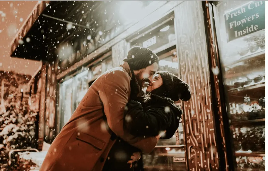 Pareja besándose bajo la nieve frente a una floristería